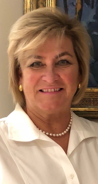 Headshot of a smiling woman with short blonde hair wearing a white blouse, pearl necklace, and gold earrings, with framed artwork in the background.