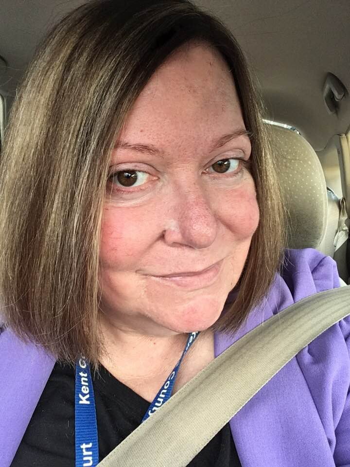 Close-up selfie of a person with short brown hair sitting in a car, wearing a purple hoodie and a lanyard, with a seatbelt across their chest.