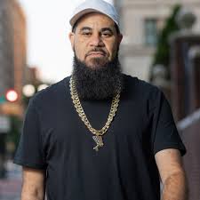 A bearded man wearing a light gray cap, a black t-shirt, and a thick gold chain necklace stands outdoors on a city street. The background is softly blurred with buildings and street elements visible.