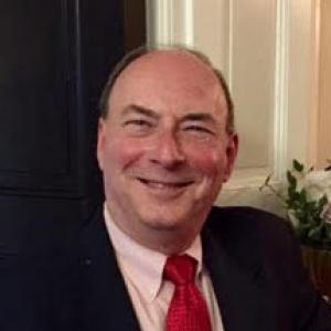Headshot of a smiling man in a dark suit jacket with a white shirt and red tie, photographed indoors.