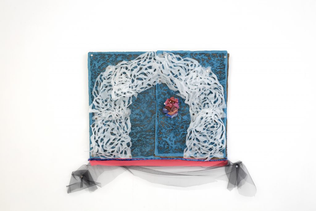 Small mixed-media wall piece with a textured turquoise-blue background and a layered white arch made of intricate cut, looping forms. A small pink and purple sculptural element sits near the center, while black tulle and a bright pink strip extend below the rectangular piece against a white wall.
