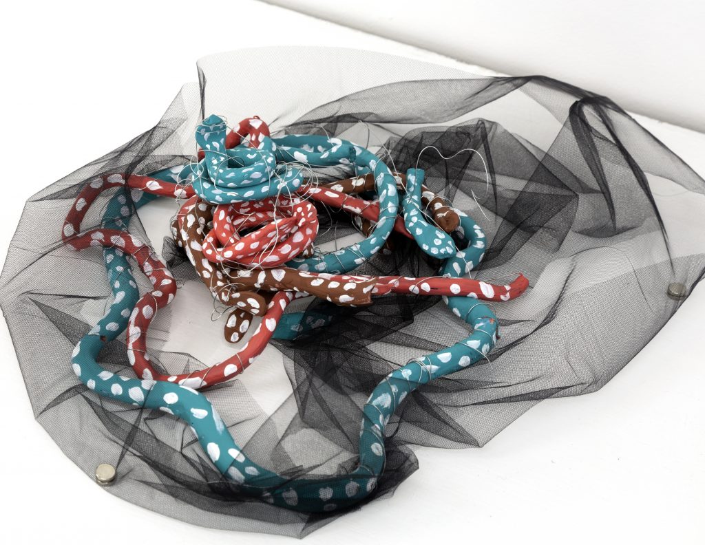 Floor-based mixed-media sculpture made of long coiled tube-like forms in turquoise, coral red, and brown, each painted with white dots. The intertwined shapes rest on black sheer fabric spread across a white floor, with thin wires looping through the sculpture and creating a nest-like arrangement.
