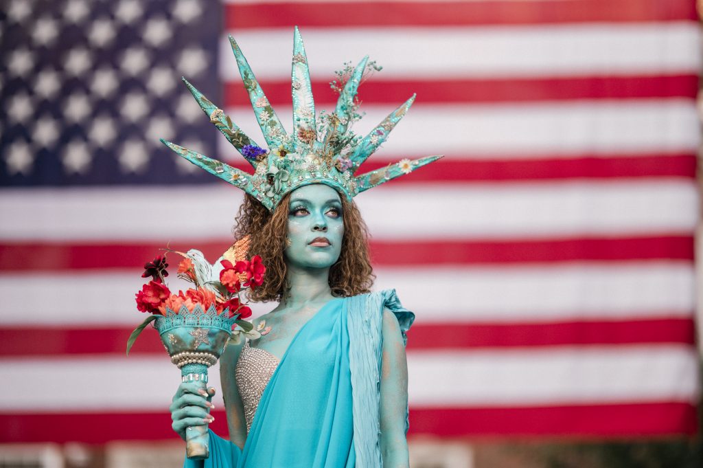 Person styled as a Statue of Liberty figure, painted turquoise and wearing an elaborate spiked crown decorated with flowers and embellishments. They hold a floral torch and stand before a blurred American flag backdrop, creating a striking performance or costume portrait.