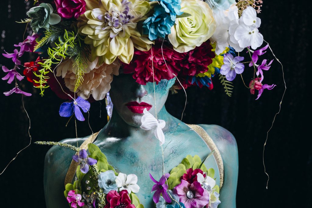 Close-up portrait of a person with turquoise body paint and bright red lipstick, wearing an oversized headdress made of large colorful flowers that drape over the eyes. Floral elements across the chest and shoulders, along with the dark background, give the image a lush, surreal, and highly stylized look.