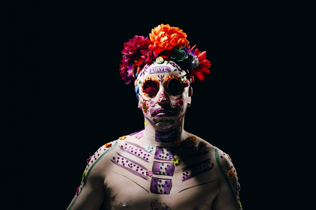 Person posed against a black background in dramatic Día de los Muertos-inspired body paint, with a flower crown and round dark glasses incorporated into the face design. Bright floral details, painted skeletal motifs, and jewel-like decorations create a vivid, theatrical portrait.