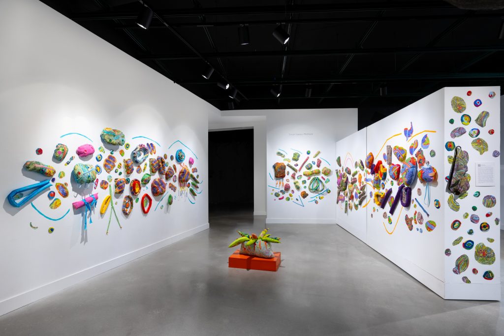 Wide gallery view of a large-scale installation featuring dozens of colorful soft sculptural forms arranged across three white walls. Brightly patterned organic shapes in pink, green, orange, blue, and purple appear to float and scatter through the space, with a small sculpture on a low orange platform placed in the center of the room.