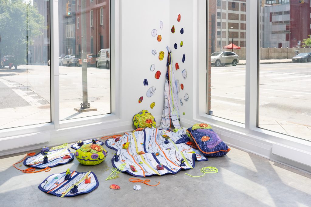 Corner installation of soft sculptural pieces spread across a concrete floor and climbing up a white wall between tall windows. The brightly colored forms and stitched fabric elements in lime green, blue, orange, and purple are connected by cords and scattered shapes, while sunlight from the street-facing windows fills the space.