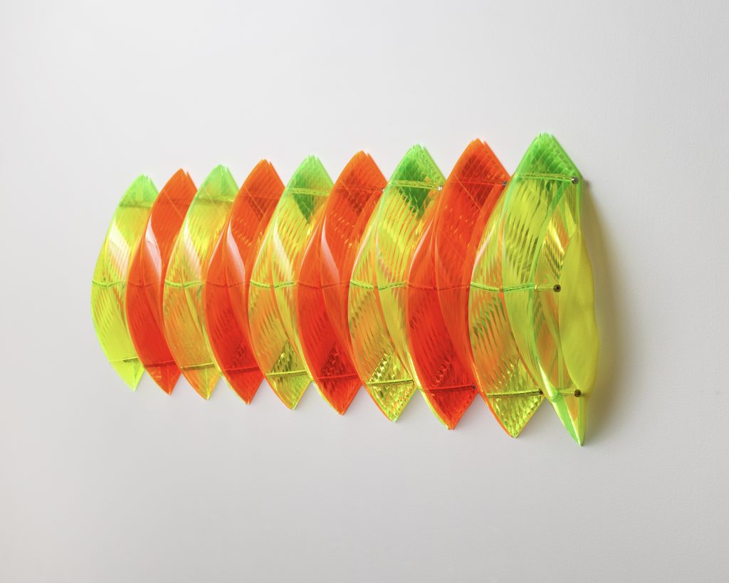 Wall-mounted abstract sculpture made of repeating translucent curved elements in fluorescent orange and yellow-green. The layered, ribbed forms create a rhythmic, woven effect against a plain white wall.