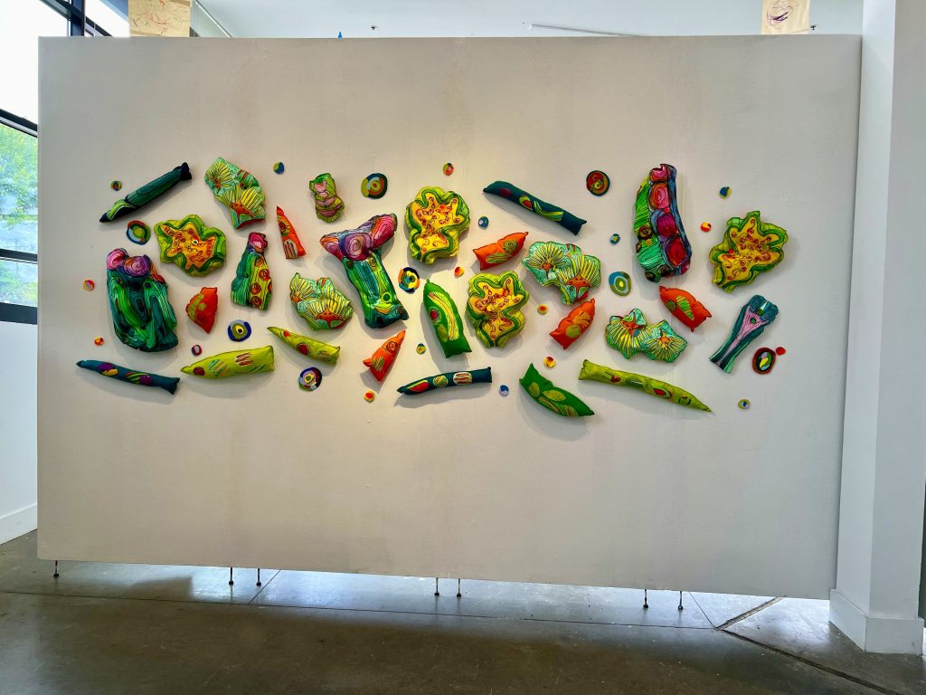 Wall-mounted installation of colorful padded organic forms arranged in a loose horizontal composition on a freestanding white panel. The shapes resemble sliced vegetables, seeds, or biological fragments, rendered in vivid greens, yellows, oranges, pinks, and blues against the plain backdrop.