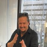 Photo of a smiling man with long hair pulled back and a goatee, wearing a black button-down shirt and sunglasses on his head, posing indoors with hands raised in a playful stance near a large window.