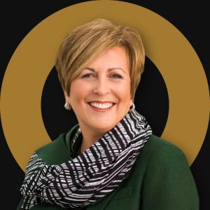 Professional headshot of a smiling woman with short blonde hair, wearing a green jacket and a black-and-white patterned scarf. A black background with a gold circular graphic frames the portrait.
