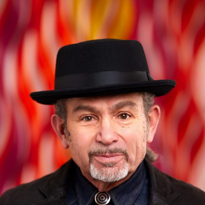 Headshot of a man in a black fedora and jacket against a red-and-orange background.
