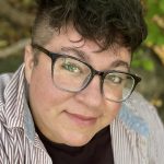 Close-up portrait of a person wearing large black glasses and an eyebrow piercing, with short curly hair, photographed outdoors.