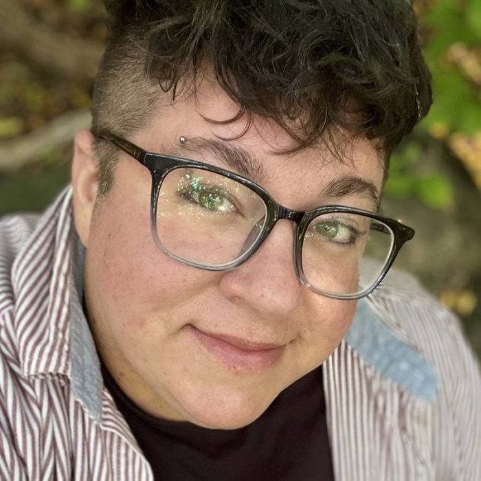 Close-up portrait of a person wearing large black glasses and an eyebrow piercing, with short curly hair, photographed outdoors.
