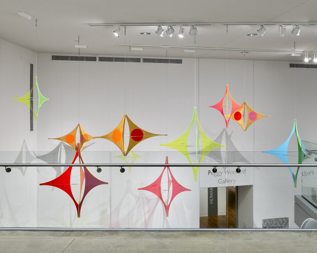 Wide view of a contemporary gallery installation featuring multiple suspended geometric sculptures in bright neon colors, including lime green, orange, pink, and blue. The translucent, kite-like forms hang at different heights above a glass railing in a white, high-ceilinged space near the Peggy Woodard Gallery entrance.