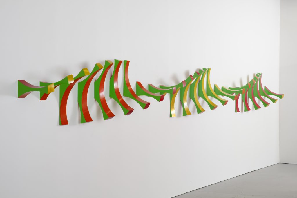 Long abstract wall sculpture composed of repeating curved shapes in green, orange, yellow, and pink. The piece stretches horizontally across a white gallery wall, creating a wave-like sense of movement and depth.