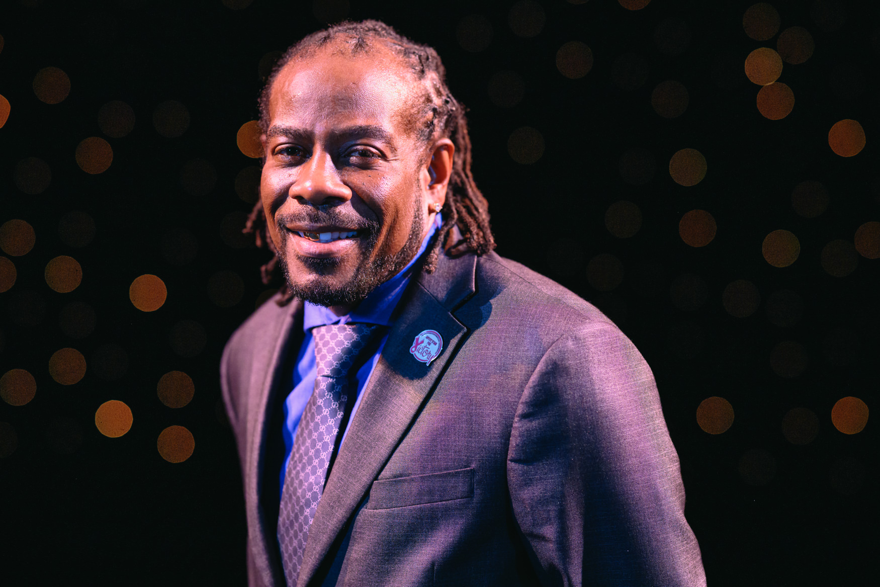 Portrait of a smiling man with short locs and a beard, wearing a dark suit, blue shirt, patterned tie, and circular lapel pin. He stands against a dark background dotted with soft golden lights, creating a warm, formal portrait.