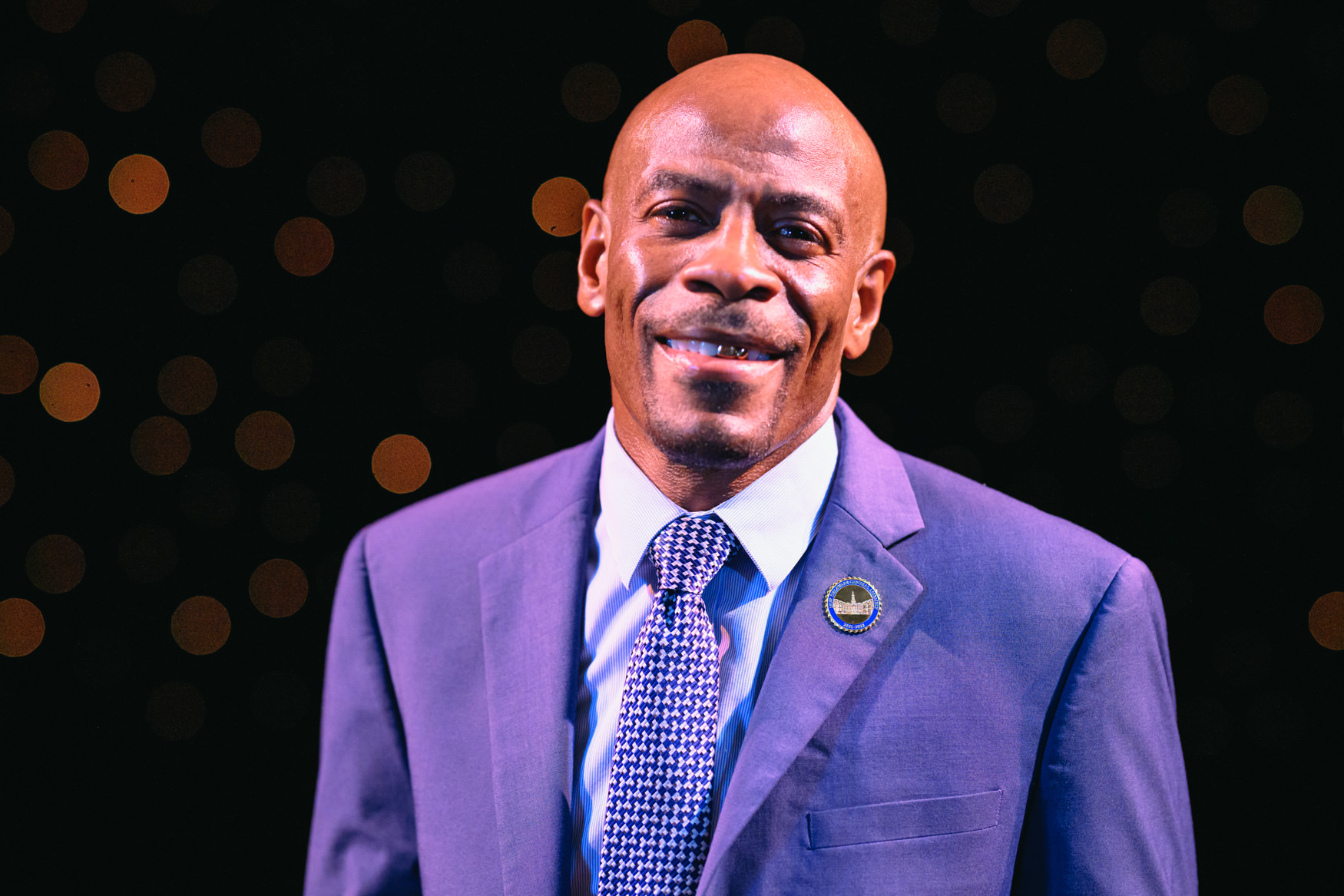 Portrait of a smiling man in a blue suit and patterned tie, wearing a circular lapel pin, against a dark background filled with soft golden lights. The close-up composition and warm stage lighting give the image a formal yet approachable feel.