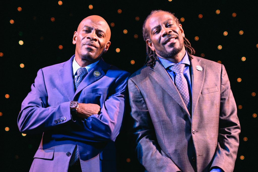 Two men in suits pose side by side against a dark background filled with soft golden lights. One stands with arms crossed and the other smiles with hands relaxed, creating a confident, formal portrait with a warm stage-lit atmosphere.