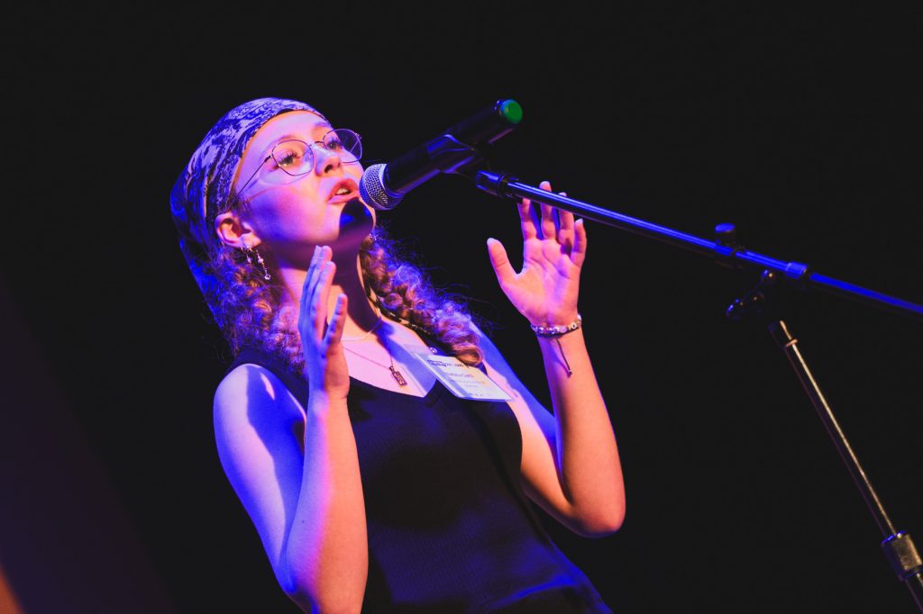 Student performer speaking or reciting into a microphone onstage under blue and purple lighting. She wears glasses, a patterned headscarf, and a black sleeveless outfit, with both hands raised expressively as she addresses the audience.