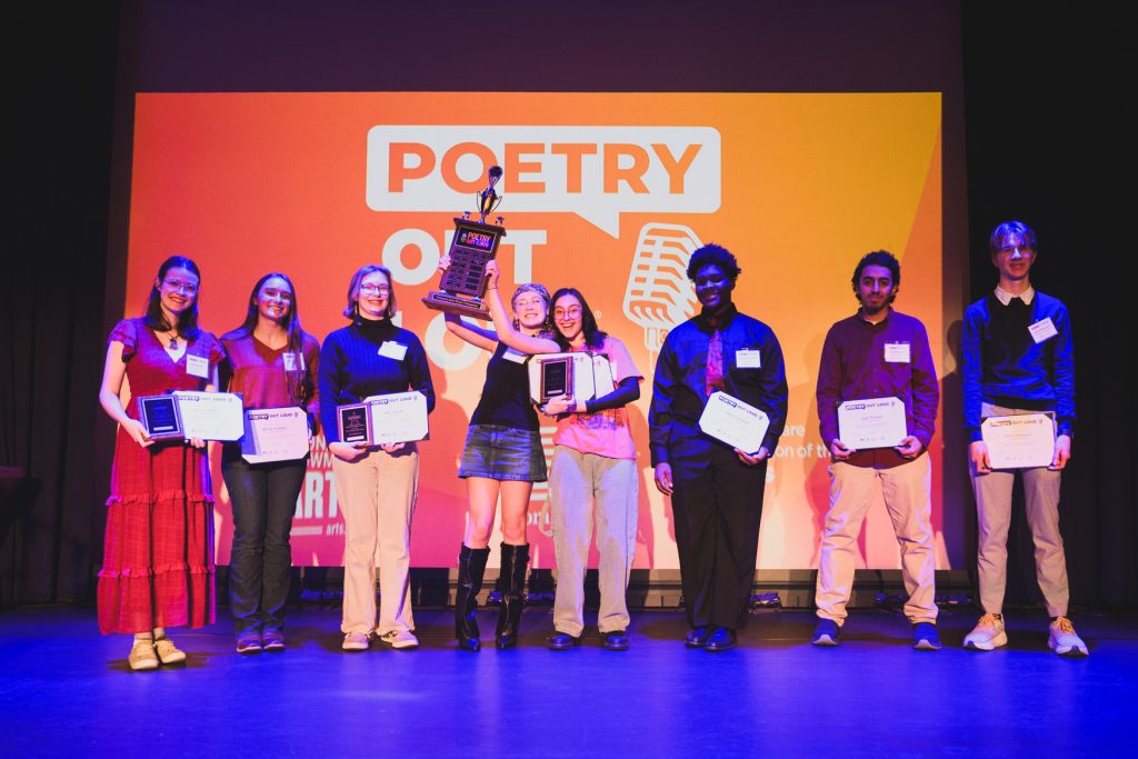 Group of student participants standing onstage with certificates and plaques in front of a large orange “Poetry Out Loud” backdrop. Two students at center hold trophies and awards overhead while the others smile and pose under stage lighting during an awards presentation.