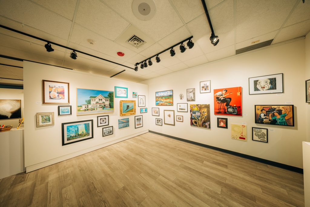 Wide view of a gallery exhibition featuring a salon-style arrangement of framed artworks hung across two white walls. The room includes paintings, drawings, and mixed-media pieces in a variety of sizes and styles, illuminated by track lighting above and set against a light wood floor.
