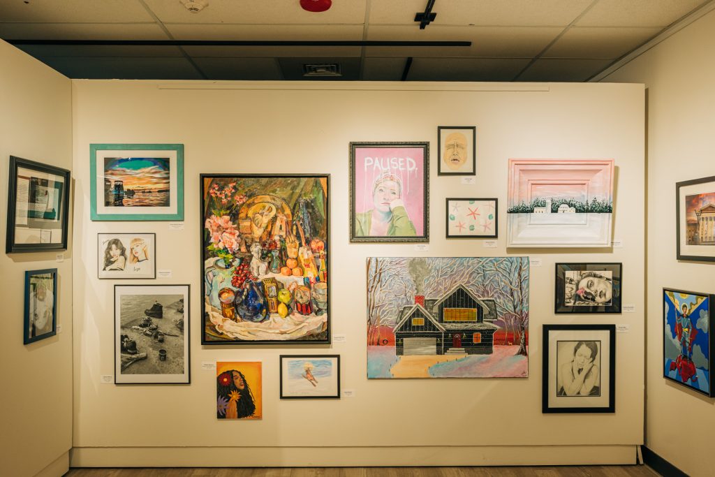 Gallery wall filled with a variety of framed artworks, including paintings, drawings, and photographs arranged salon-style against a white partition. The display features landscapes, portraits, a colorful still life, and a winter house scene, all lit by overhead track lighting in an exhibition setting.
