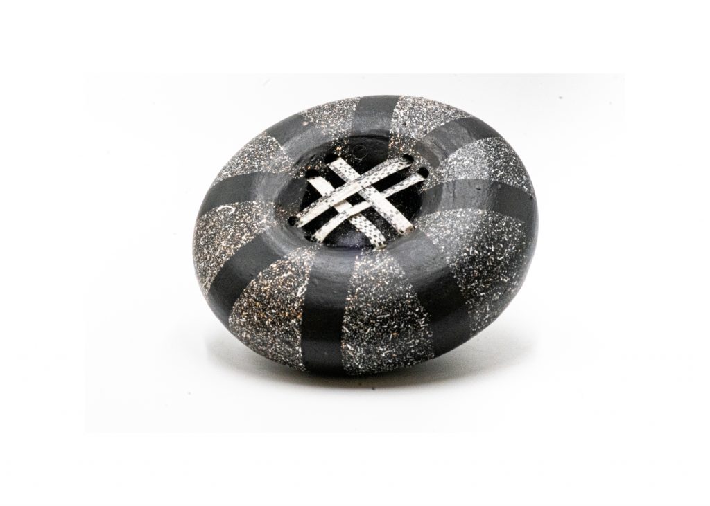 Flattened round ceramic vessel with a black-and-silver speckled surface patterned in alternating dark curved bands. The recessed opening at the center is crossed by two narrow white strips, giving the piece a woven or lattice-like detail.
