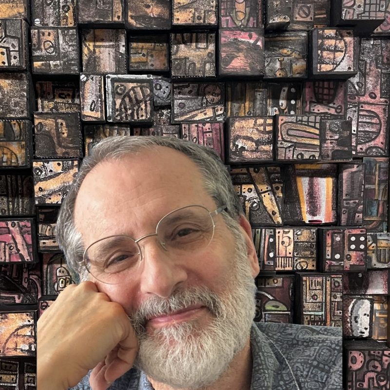 Smiling man with a gray beard and round glasses in front of an abstract patterned wall.