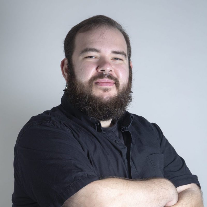 Studio portrait of a bearded person with a septum ring wearing a black button-down shirt, arms crossed, against a light gray background.