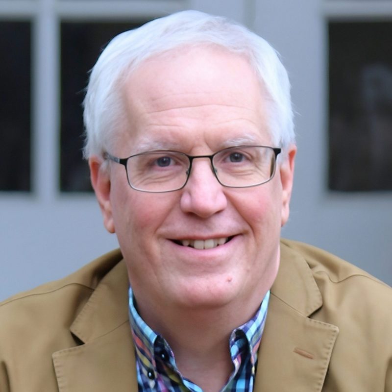 Headshot of a smiling man with white hair and glasses, wearing a tan blazer over a multicolored plaid shirt, with a softly blurred background.