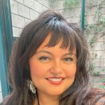 Close-up portrait of a smiling woman with long dark hair and bangs, wearing a black top and a septum ring, photographed outdoors near a brick wall with climbing greenery.