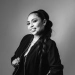 Black-and-white portrait of a woman posing with a slight smile against a plain background. She wears a dark zip-front jacket, hoop earrings, and a long styled ponytail, creating a polished, confident studio portrait.
