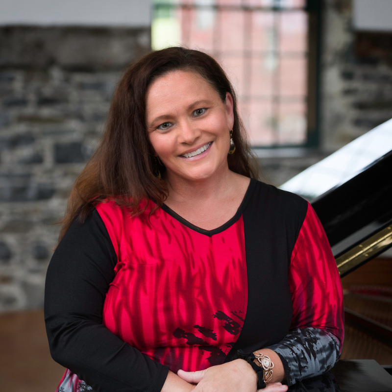 Portrait of a smiling woman with long brown hair, wearing a red-and-black patterned top and standing beside a grand piano. The softly blurred stone wall and window behind her create a warm, elegant indoor setting.