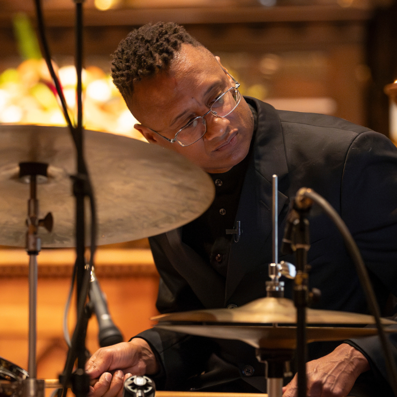 Portrait of a musician seated at a drum set, wearing glasses and a black suit while looking down in concentration. Warm golden light and softly blurred background details give the image an intimate performance atmosphere.