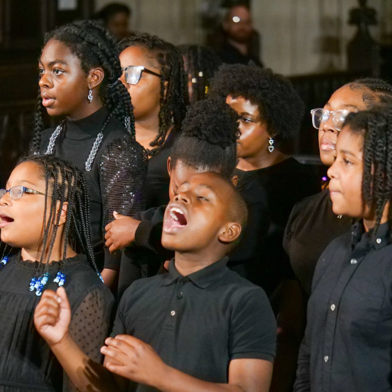 Group of children dressed in black performing together, singing with expressive faces and focused energy. The close-up composition highlights several young vocalists, with one child in the center singing passionately and others gathered closely around.