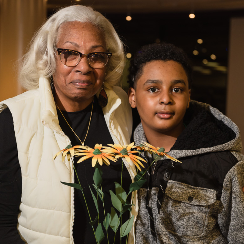 Portrait of an older woman with white hair and glasses standing beside a young boy in a gray-and-black jacket. They pose together indoors, holding decorative yellow flowers, with warm lighting and a softly blurred background.
