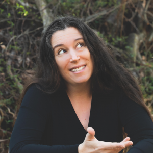 Portrait of a smiling woman with long dark hair, wearing a black top and seated outdoors against a natural background of branches and greenery. She looks upward with an expressive, conversational gesture, giving the image a warm and candid feel.