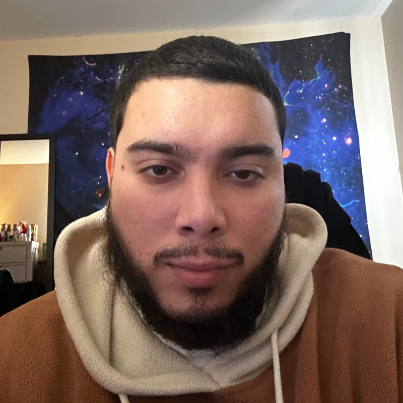 Close-up portrait of a young man with short dark hair and a trimmed beard, looking directly at the camera with a neutral expression. He wears a tan fleece hoodie layered over a cream-colored hood, and a dark blue galaxy-pattern tapestry hangs on the wall behind him.