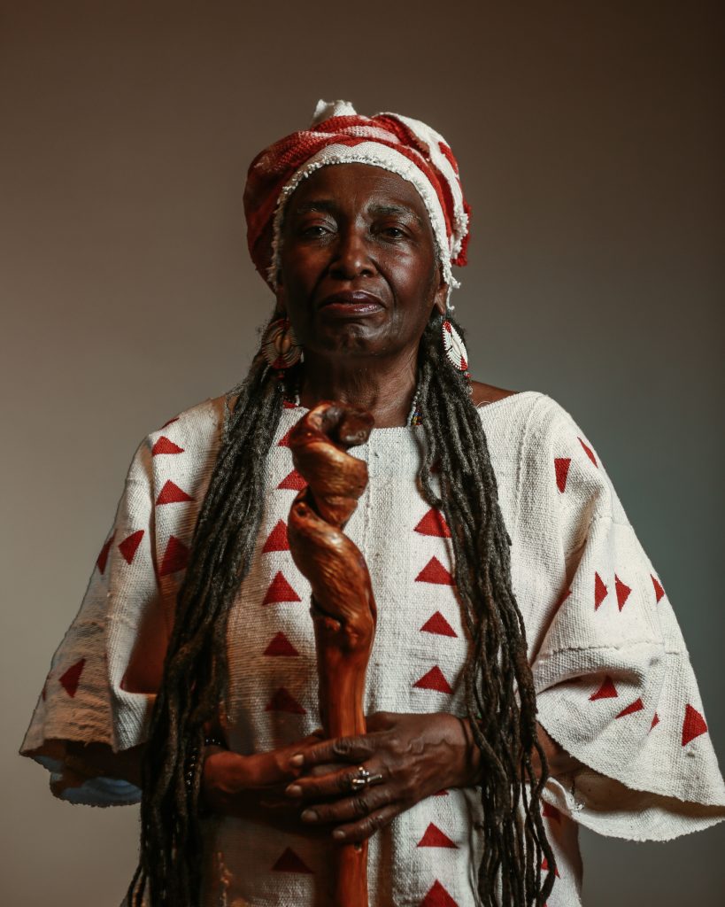 Studio portrait of an older woman standing against a neutral background, wearing a white woven garment and matching headwrap patterned with red triangles. She holds a carved wooden staff in both hands, and her long gray locs and beaded earrings frame a calm, dignified expression.