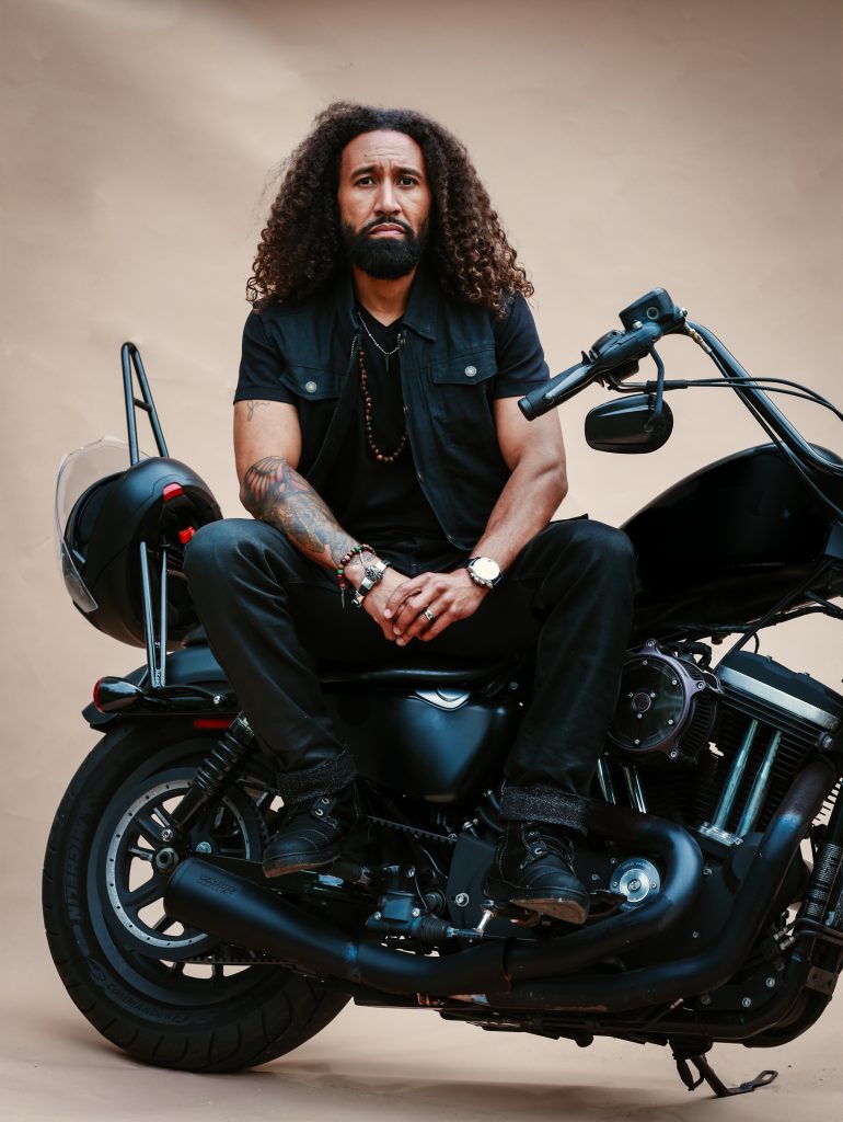 Studio portrait of a man seated on a black motorcycle against a plain beige backdrop. He wears an all-black outfit with boots, bracelets, and a watch, and his long curly hair and direct gaze give the image a strong, self-possessed presence.