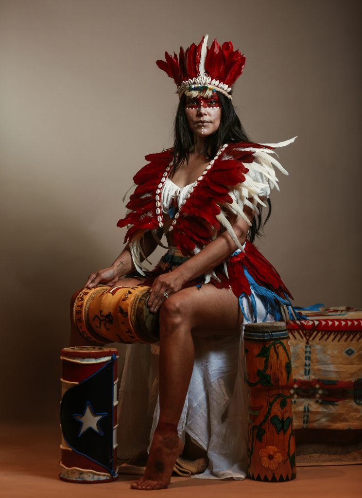 Studio portrait of a woman seated barefoot in ceremonial dress, wearing a dramatic red-and-white feathered headdress and matching feathered shoulder adornments. She holds a decorated drum across her lap, with painted drums and patterned textiles around her, and faces the camera with a poised, confident expression.