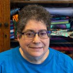 Headshot of a person with short curly hair wearing blue-framed glasses and a bright blue shirt, photographed indoors in front of shelves of colorful folded fabric.