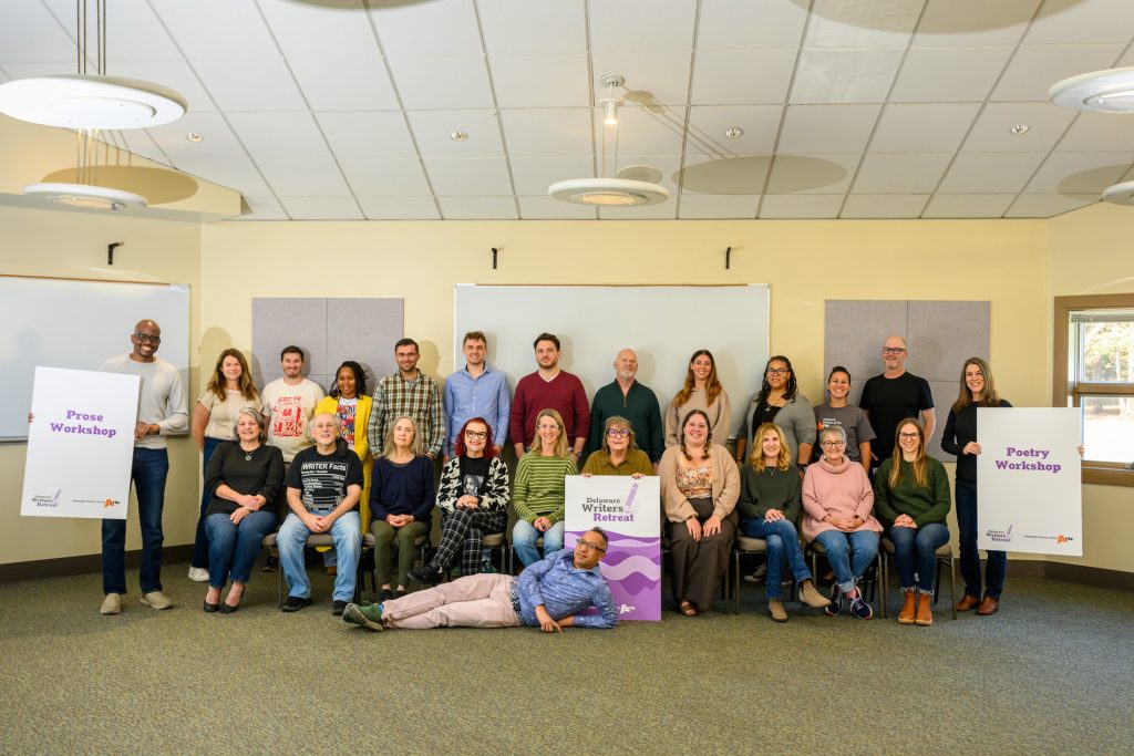 Large group photo of participants and facilitators at the Delaware Writers Retreat, posed in a meeting room with workshop signs for prose and poetry. Two people hold large signs at either side, another person reclines playfully at the front, and the group smiles around a central Delaware Writers Retreat sign.