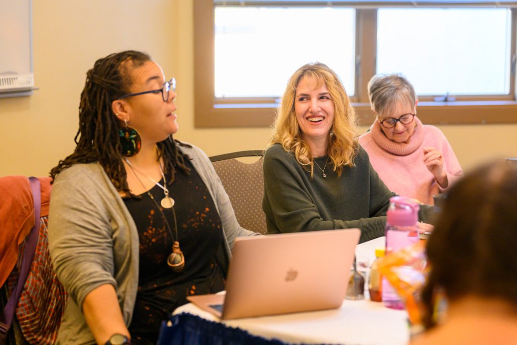 Three women sit at a table engaged in conversation.  The woman on the left is speaking and has a laptop in front of her.  The middle woman is looking at the speaker and smiling.  The third woman is smiling and looking down.