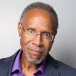 Headshot of a man with short gray hair and glasses, wearing a navy blazer over a purple shirt, smiling gently against a light gray background.