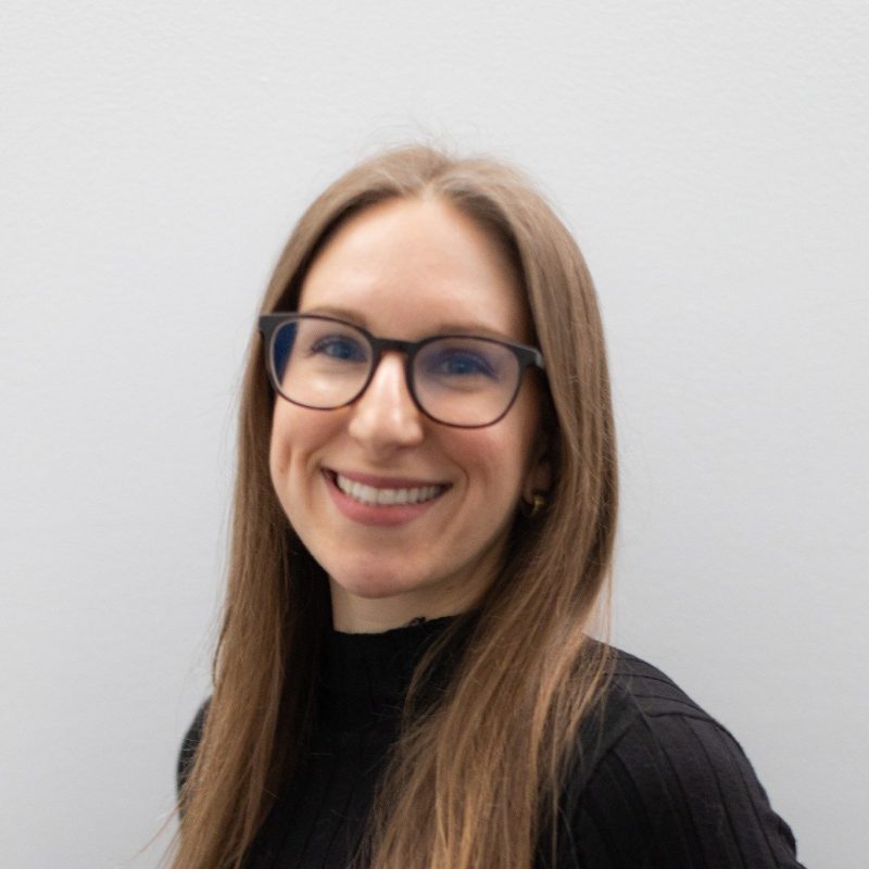 Portrait of a smiling woman with long light-brown hair wearing glasses and a black top, photographed against a light gray background.