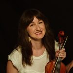 Portrait of a smiling woman with long brown hair holding a violin and bow, wearing a light top and blue skirt against a dark background.