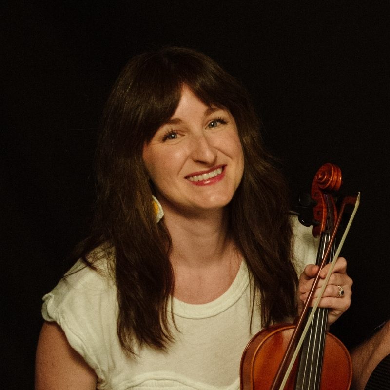 Portrait of a smiling woman with long brown hair holding a violin and bow, wearing a light top and blue skirt against a dark background.