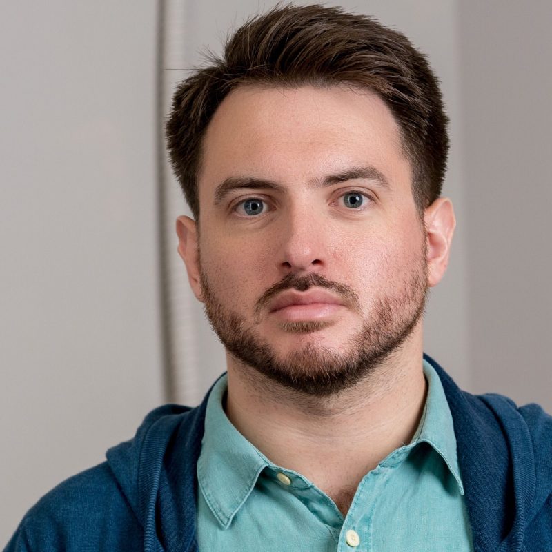 Headshot of a man with short dark hair and a trimmed beard, wearing a teal button-down shirt and a blue hoodie, looking at the camera against a neutral indoor background.